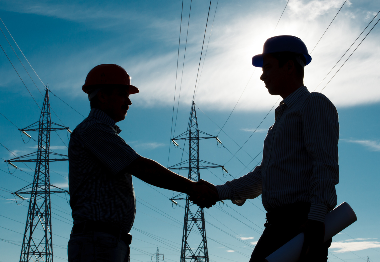 Two workers wearing hard hats shaking hands infront of two electrical transmission power lines.