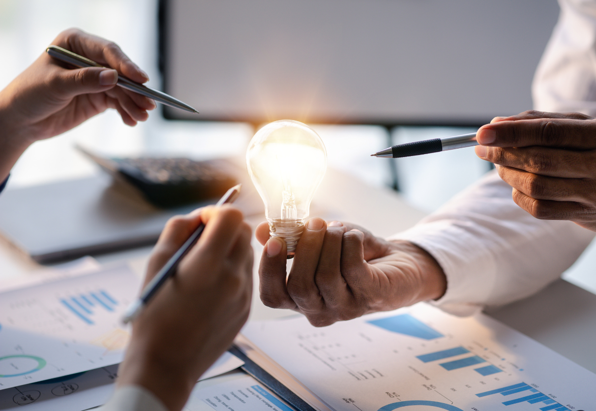 Three persons pointing at a shining lightbulb with pens among a desk with charts on it.